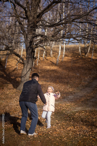 Loving father playing with little daughter both having fun and smiling having walk in forest with dry fallen leaves on ground on sunny fall day. Family time together.