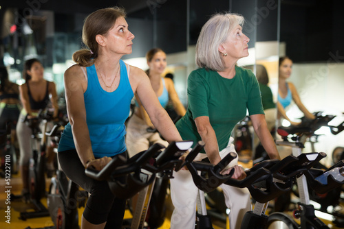 Two mature women doing cardio workout at gym, training together on exercise bikes