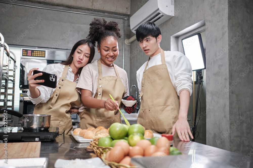 Fototapeta premium Three young students in cooking class wear aprons enjoyment while taking selfie photo with mobile phone in kitchen, smiling and laughing, preparing eggs and fruits, learn fun culinary course together.