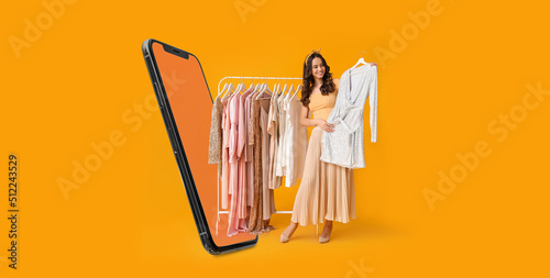 Stylish young woman with stylish clothes on hanger and modern mobile phone on yellow background photo