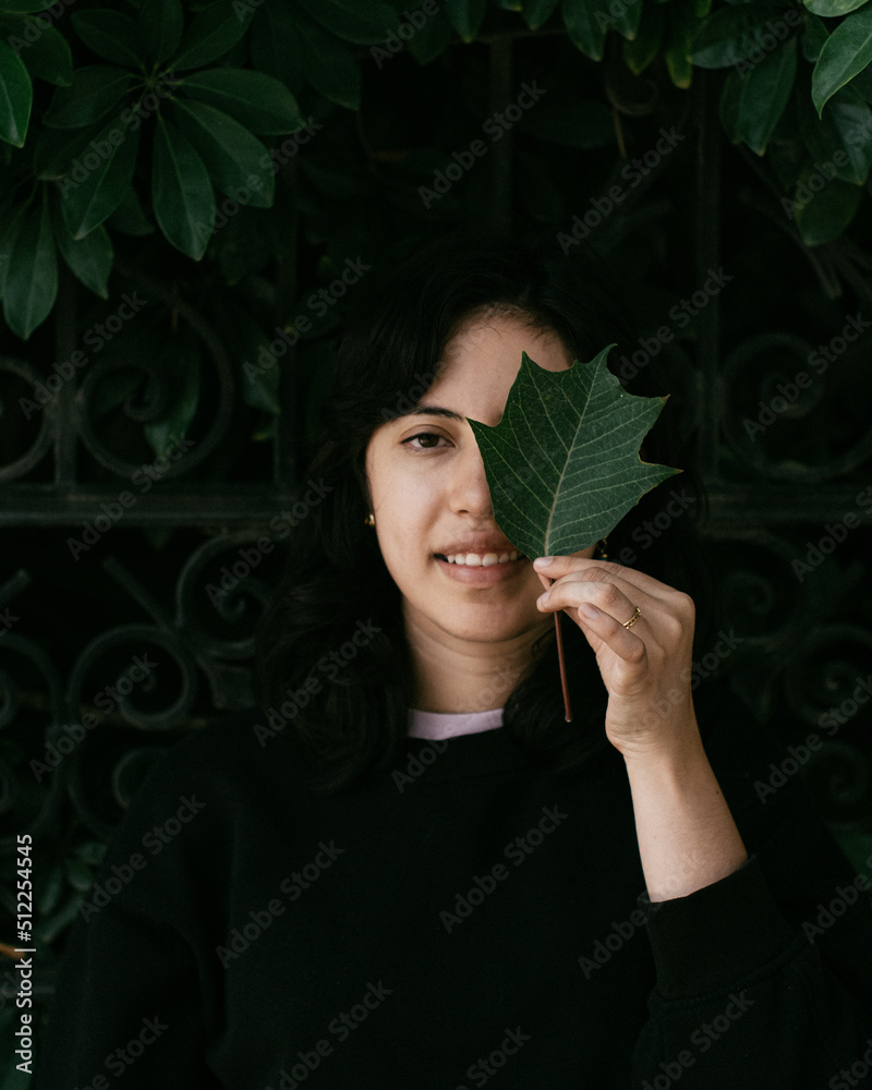 Obraz premium photo of a cheerful young woman dressed in black living around nature in a joyful way holding a leaf near her eye with a background of a fence with black ironwork and many plants