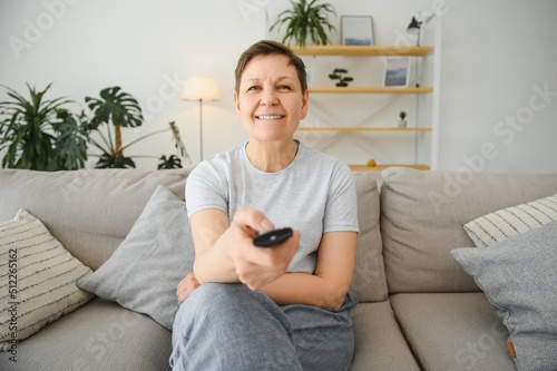 middle-aged woman sitting on the sofa and turning on the TV with a remote control.
