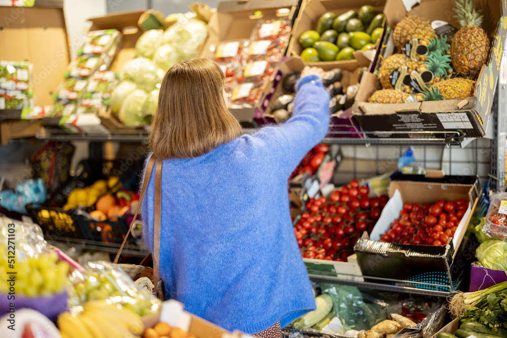Obraz premium Woman choosing avocado from the counter full of products at market. View from the backside. Buying food at local market