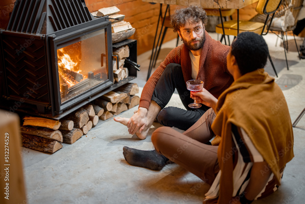 Two men talking while sitting together by the burning fireplace at cozy home. Concept of homosexual relations and coziness on winter time. Idea of multinational gay families