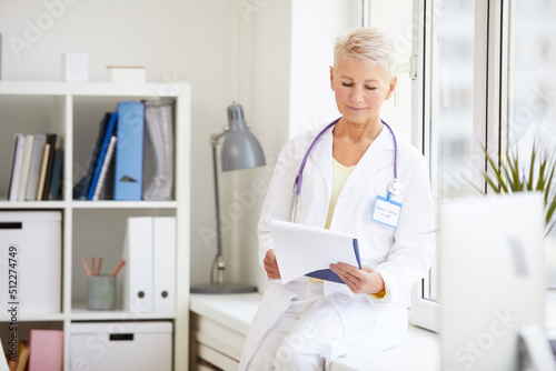Smiling attractive mature doctor in lab coat sitting on window-sill and reading patients medical record in clipboard