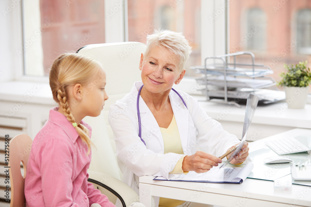 Fototapeta premium Content friendly mature female pediatrician pointing at x-ray image while explaining reason of pain to little girl in childrens healthcare clinic