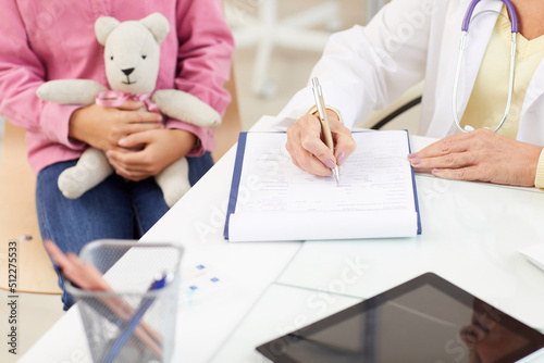 Close-up of unrecognizable doctor sitting at table and filling document while talking to youthful patient during medical consultation