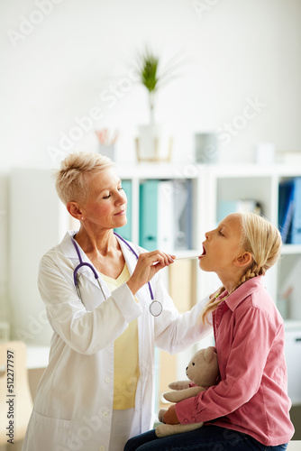 Serious attractive therapist in lab coat using medical spatula while checking throat of child with toy