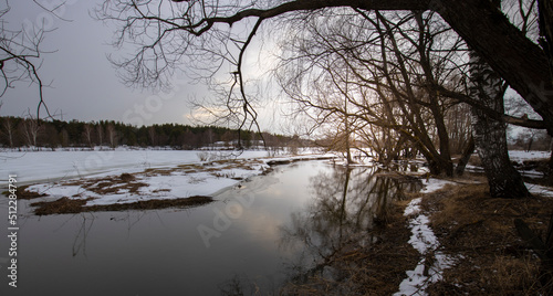 Epic spring landscape by the river. March evening, snow melts. Winding river with snow-covered banks and ice. Trees are reflected in the water.