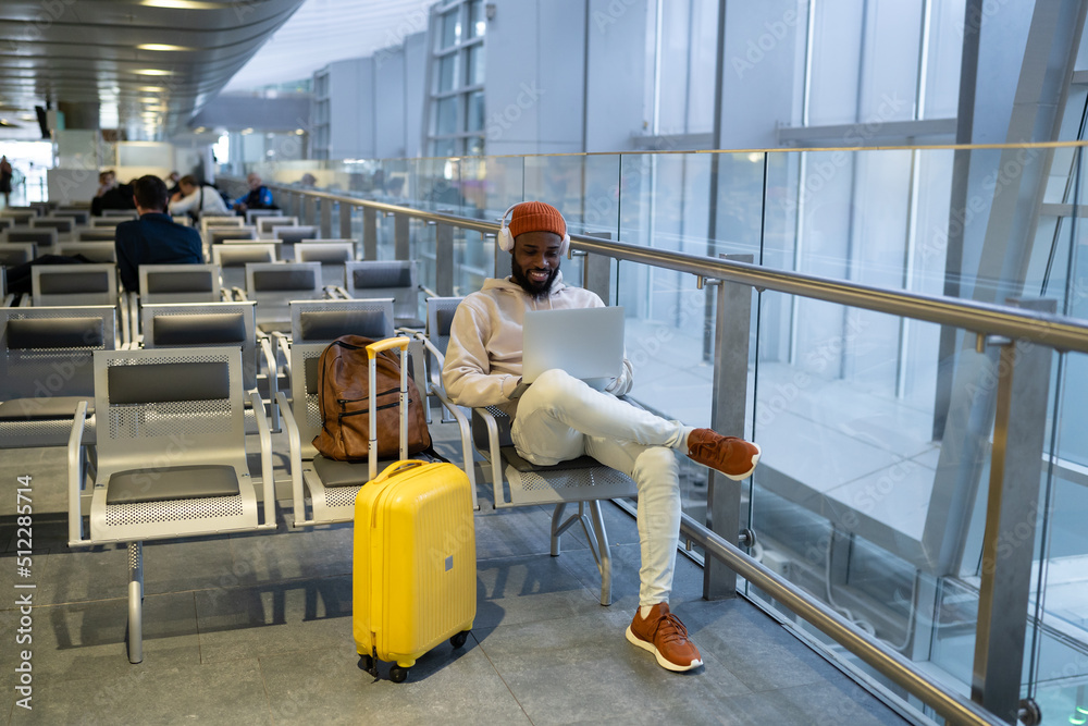 Smiling African American man sitting in hall of airport terminal using laptop, wear headphones and listening music. Passenger travelling with backpack, online working on computer and waiting landing. 