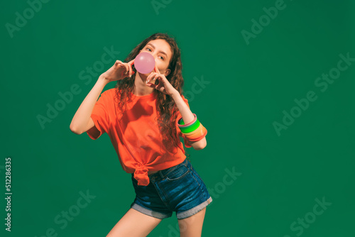 Portrait of cheerful young girl in casual summer outfit, posing with bubble gum isolated over green studio background