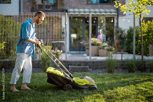 Man mows the lawn with lawn mower at backyard of his house. Husband takes care of garden on summer evening. Modern electric wireless garden equipment photo