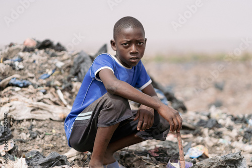 Young African waste picker boy sitting crouched in a landfill surrounded by plastic garbage, looking at the camera with a frown; symbol of stolen childhood and child labour photo