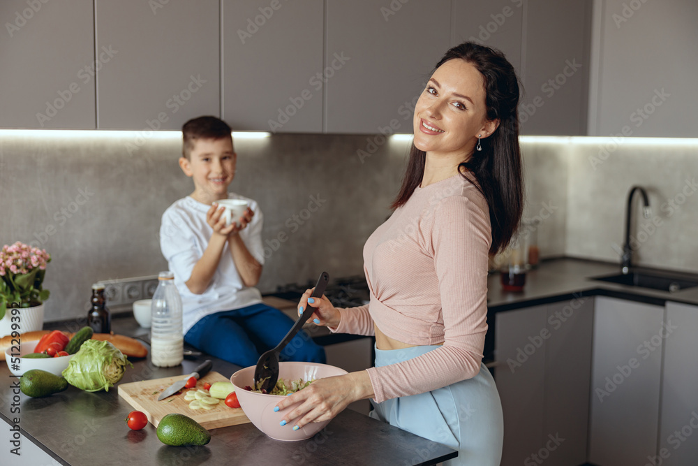Portrait of beautiful Caucasian young mother cooking salat in kitchen. Small son on background. Portrait of beautiful Caucasian young mother cooking salat in kitchen. Small son on background.