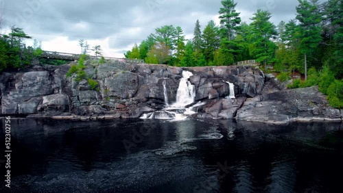 Rushing waterfall cascades through natural bedrock cliff in Northern Ontario with forest and cloudy sky photo