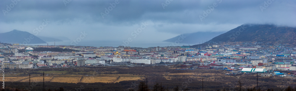 Panorama of the city of Magadan. Top view of a seaside town located among the mountains on the coast of a sea bay. Cloudy rainy weather in early summer. Magadan region, Siberia, Far East of Russia.