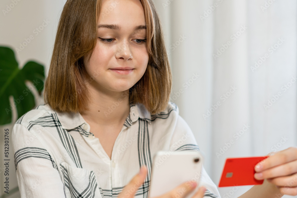 Female professional doing online payment through credit card while sitting at home office. Cheerful young girl shopping or banking online using mobile phone and credit card, living room interior. Female professional doing online payment through credit card while sitting at home office. Cheerful young girl shopping or banking online using mobile phone and credit card, living room interior.