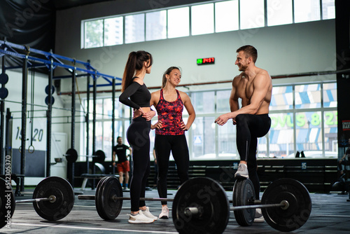 Athletic sport male athlete communicates with fit girls after training in hardcore gym. Fitness healthy man and woman tired after strength training exercise at gym.