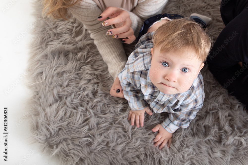 A one-year-old blond baby crawls on the carpet, surrounded by parents, looks at the camera. Top view