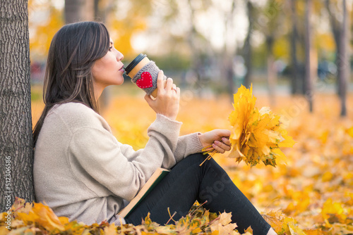 Young woman drinking coffee from paper cup with a book on her lap and an autumn yellow bouquet of leaves in an autumn park.