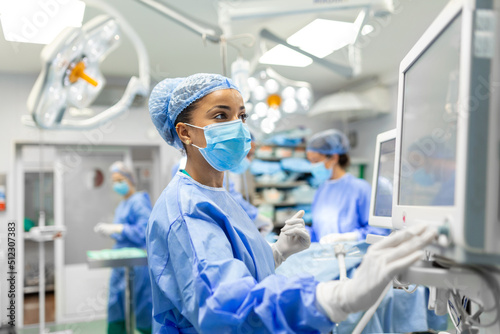 Anesthetist Working In Operating Theatre Wearing Protecive Gear checking monitors while sedating patient before surgical procedure in hospital photo