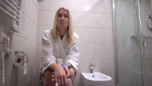 Young beautiful Caucasian woman blonde in bathrobe sits on toilet, in modern home bathroom. She thinks pensively and looks into ode of point without any emotion. Morning emptying.