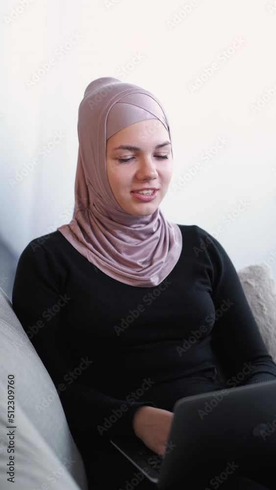 Online call. Webinar conference. Work from home. Cheerful friendly woman coach in hijab talking on internet virtual meeting at laptop indoors on couch vertical. Online call. Webinar conference. Work from home. Cheerful friendly woman coach in hijab talking on internet virtual meeting at laptop indoors on couch vertical.