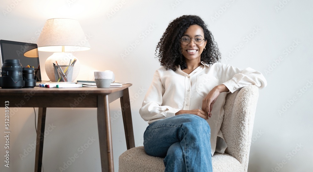 Young African American woman looking at camera and smiling. Brunette with curly hair sits at table in light home office Young African American woman looking at camera and smiling. Brunette with curly hair sits at table in light home office