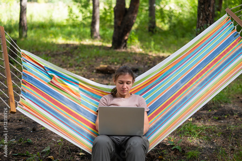 Caucasian woman working on laptop while sitting in a hammock in the forest. Girl uses a wireless computer on a hike.