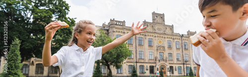 Cheerful schoolkid holding sandwich and waving hand near asian firend in park, banner. photo