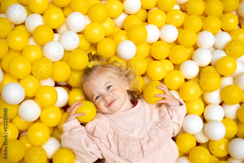 Funny cute little caucaisian blonde baby girl,toddler, smiling kid having fun in ball pool,playing with yellow white balls.Happy child looking at camera in playground,top view