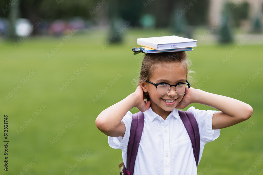 Smiling schoolgirl in eyeglasses holding notebooks on head outdoors.