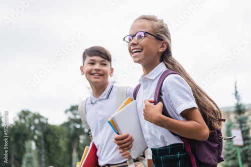 Positive schoolkid with backpack and notebooks walking near blurred asian friend outdoors.