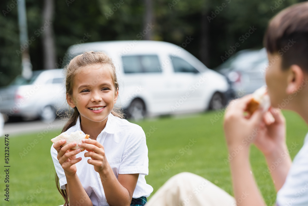 Smiling schoolgirl holding sandwich near blurred classmate in park. Smiling schoolgirl holding sandwich near blurred classmate in park.