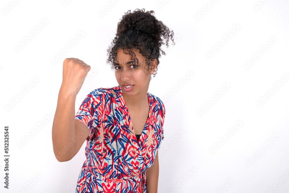 Fierce Young African American woman wearing colourful dress over white wall holding fist in front as if is ready for fight or challenge, screaming and having aggressive expression on face. Fierce Young African American woman wearing colourful dress over white wall holding fist in front as if is ready for fight or challenge, screaming and having aggressive expression on face.