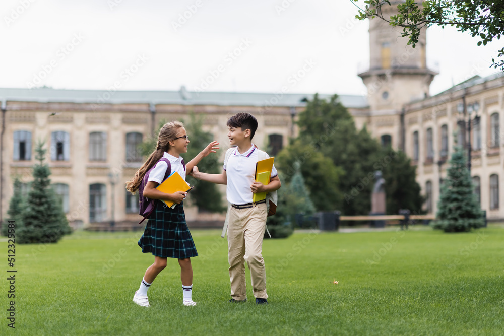 Positive multiethnic kids with backpacks and notebooks walking on grass outdoors.