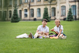 Cheerful interracial classmates holding sandwiches near lunchboxes on grass in park.