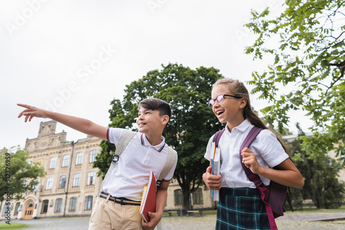 Asian schoolboy with notebooks pointing with finger near classmate outdoors. © LIGHTFIELD STUDIOS