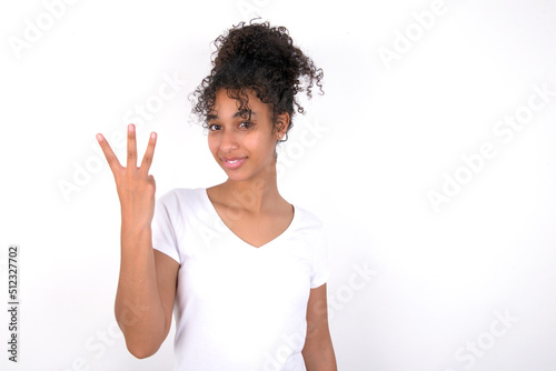 Young beautiful girl with afro hairstyle wearing white t-shirt over white wall smiling and looking friendly, showing number three or third with hand forward, counting down