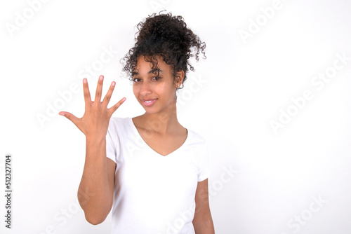 Young beautiful girl with afro hairstyle wearing white t-shirt over white wall smiling and looking friendly, showing number five or fifth with hand forward, counting down