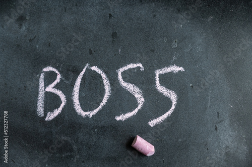 Boss. A word written in pink chalk on a black chalkboard. Handwritten text. A piece of colored chalk hangs next to it.