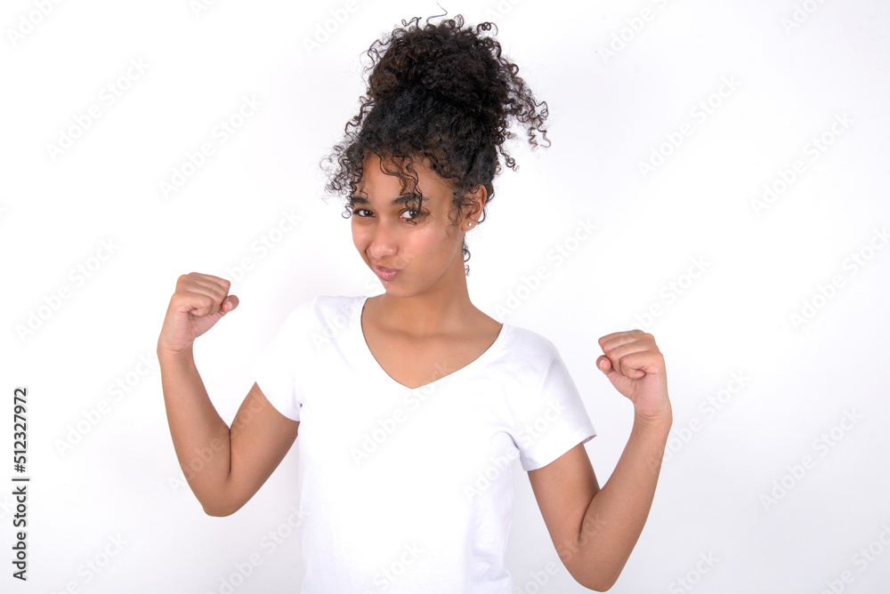 Irritated Young beautiful girl with afro hairstyle wearing white t-shirt over white wall blows cheeks with anger and raises clenched fists expresses rage and aggressive emotions. Furious model Irritated Young beautiful girl with afro hairstyle wearing white t-shirt over white wall blows cheeks with anger and raises clenched fists expresses rage and aggressive emotions. Furious model