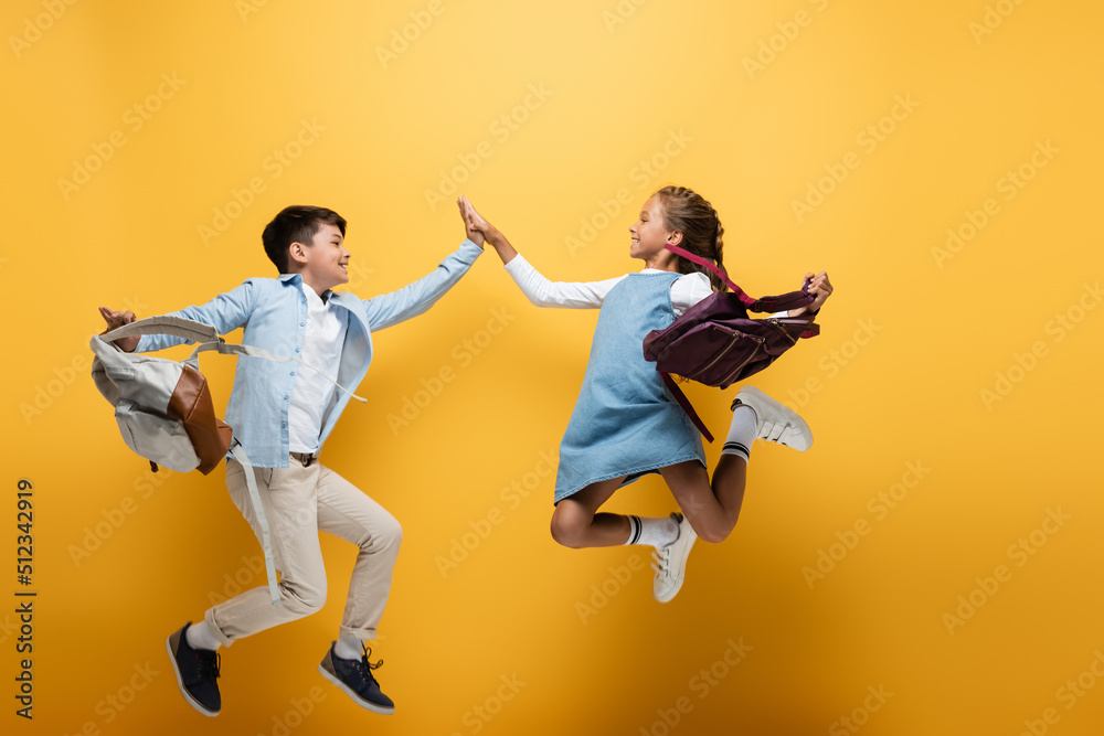 Side view of multiethnic schoolkids with backpacks giving high five while jumping on yellow background. Side view of multiethnic schoolkids with backpacks giving high five while jumping on yellow background.