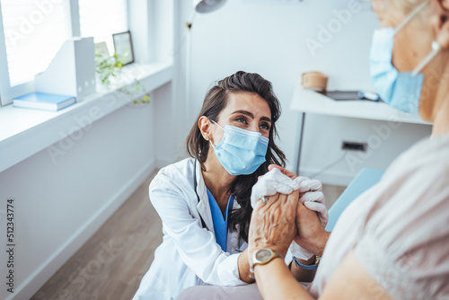 Elderly woman talking with a doctor while holding hands at home and wearing face protective mask. Worried senior woman talking to her general pratictioner visiting her at home during virus epidemic.
