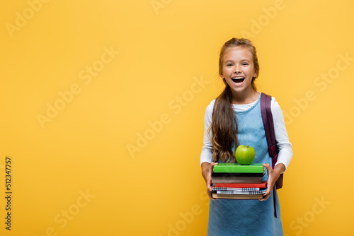 Фотография Cheerful pupil holding books and apple isolated on yellow.