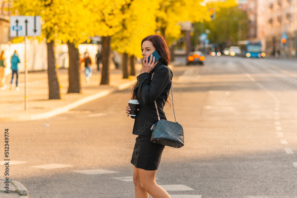 Young business woman talking on the phone holding coffee paper cup,crossing street blurred background. Fashion business photo of beautiful girl in black suite with phone and cup of coffee.Side view.