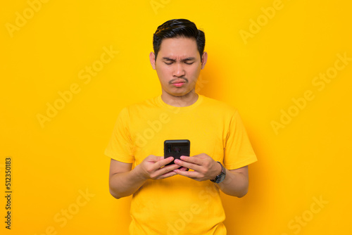 Serious young Asian man in casual t-shirt using mobile phone and looking at gadget screen isolated on yellow background. People lifestyle concept © Sewupari Studio