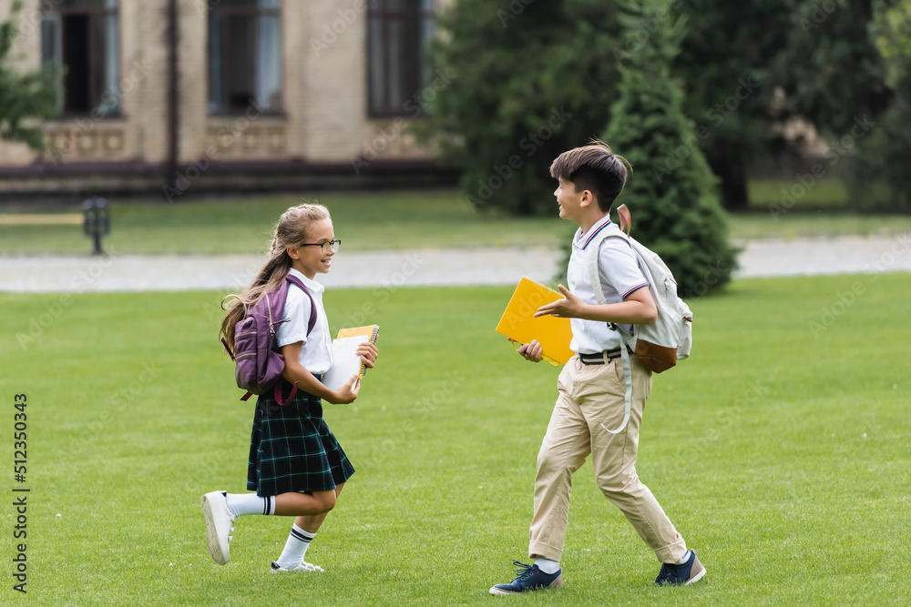Obraz premium Smiling multiethnic classmates with notebooks running on lawn in park.