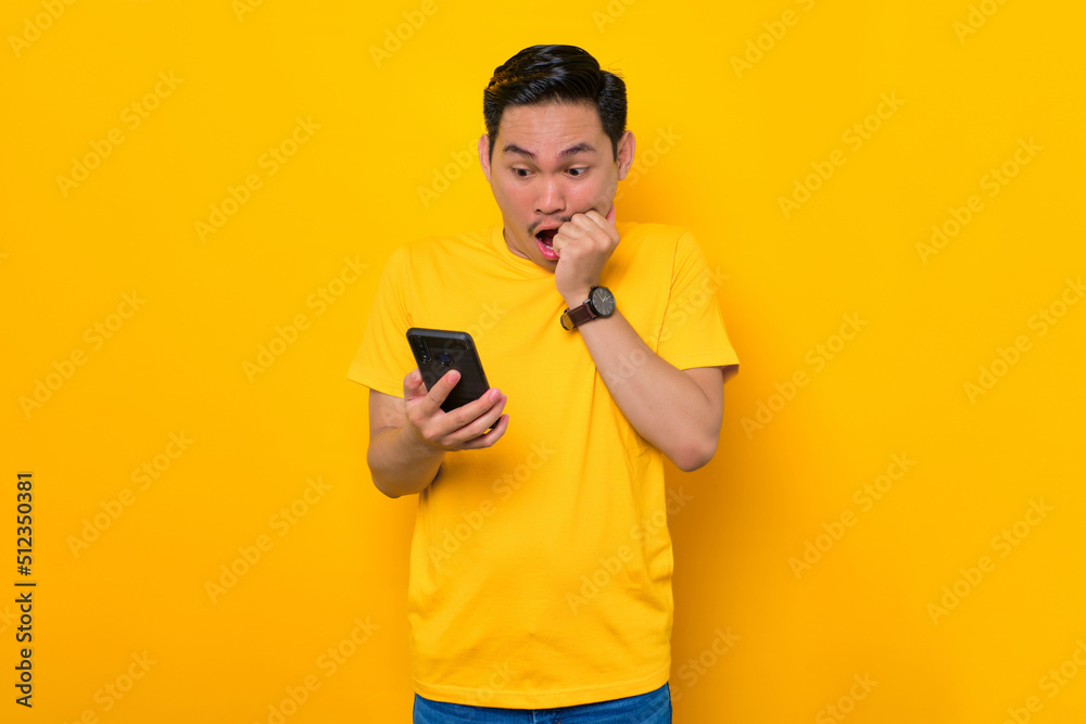 Scared young Asian man in casual t-shirt looking at mobile phone screen, reacting to bad news isolated on yellow background. People lifestyle concept Scared young Asian man in casual t-shirt looking at mobile phone screen, reacting to bad news isolated on yellow background. People lifestyle concept