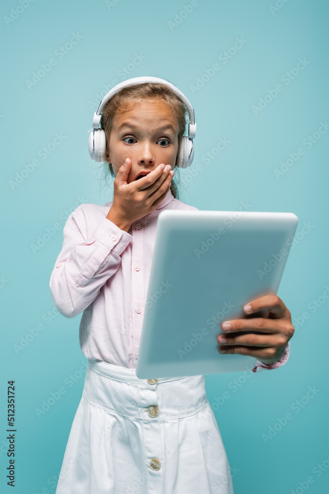 Shocked schoolgirl in headphones looking at digital tablet isolated on blue. Shocked schoolgirl in headphones looking at digital tablet isolated on blue.